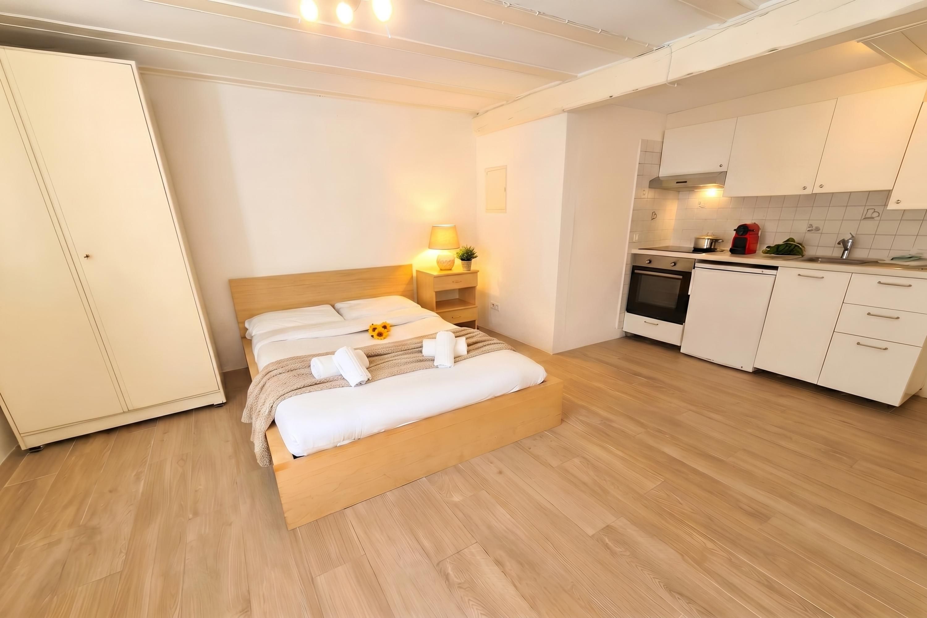 A bedroom with a wooden bed, a nightstand with a lamp, a kitchen with white cabinets, and a wooden floor.