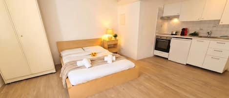 A bedroom with a wooden bed, a nightstand with a lamp, a kitchen with white cabinets, and a wooden floor.