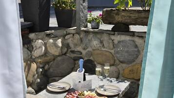 A stone wall, a table with a bottle of champagne, glasses, plates, cutlery, and a charcuterie board with meats and olives.