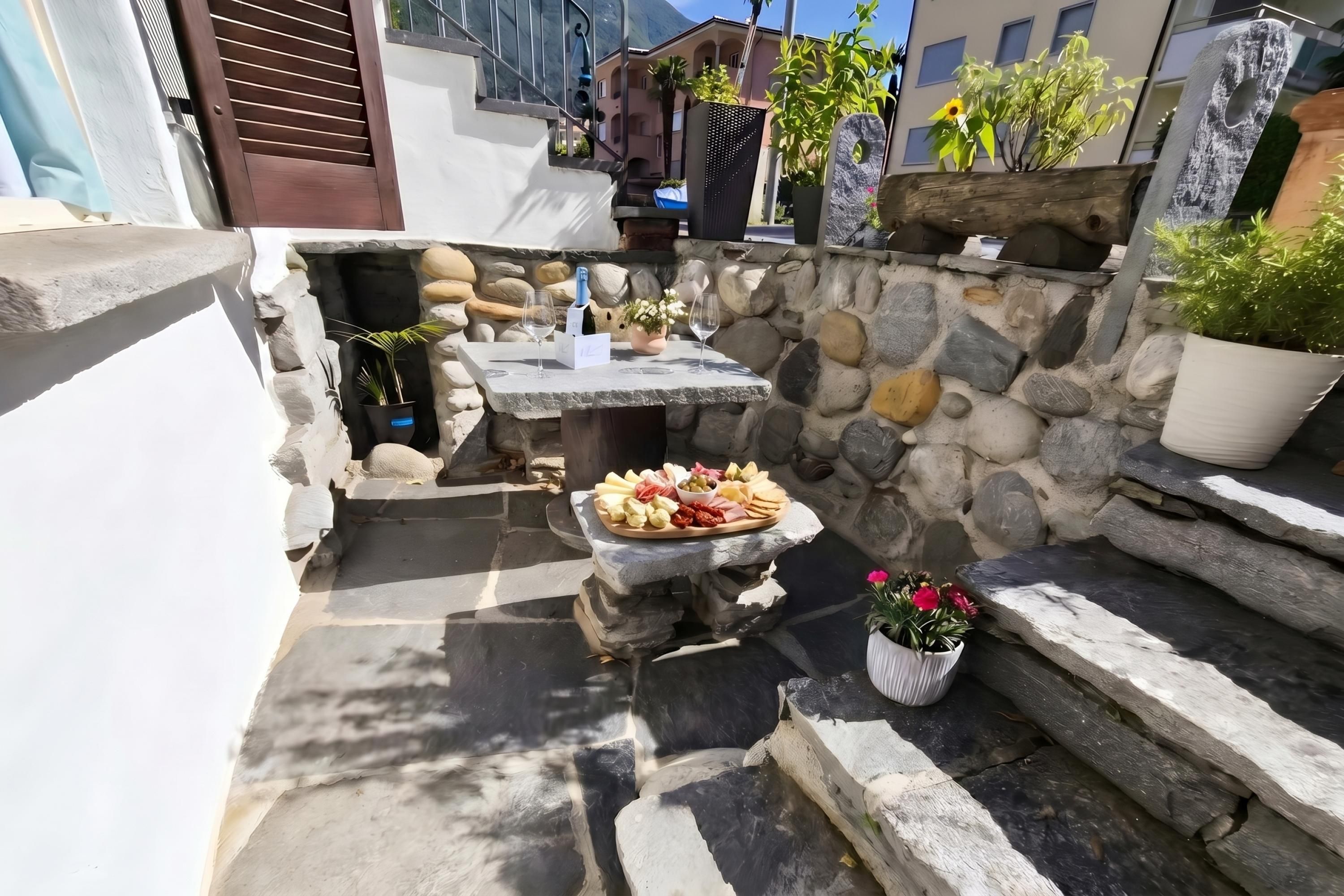 Un área de estar al aire libre con construcción de piedra, una mesa, plantas en macetas y un muro de piedra.