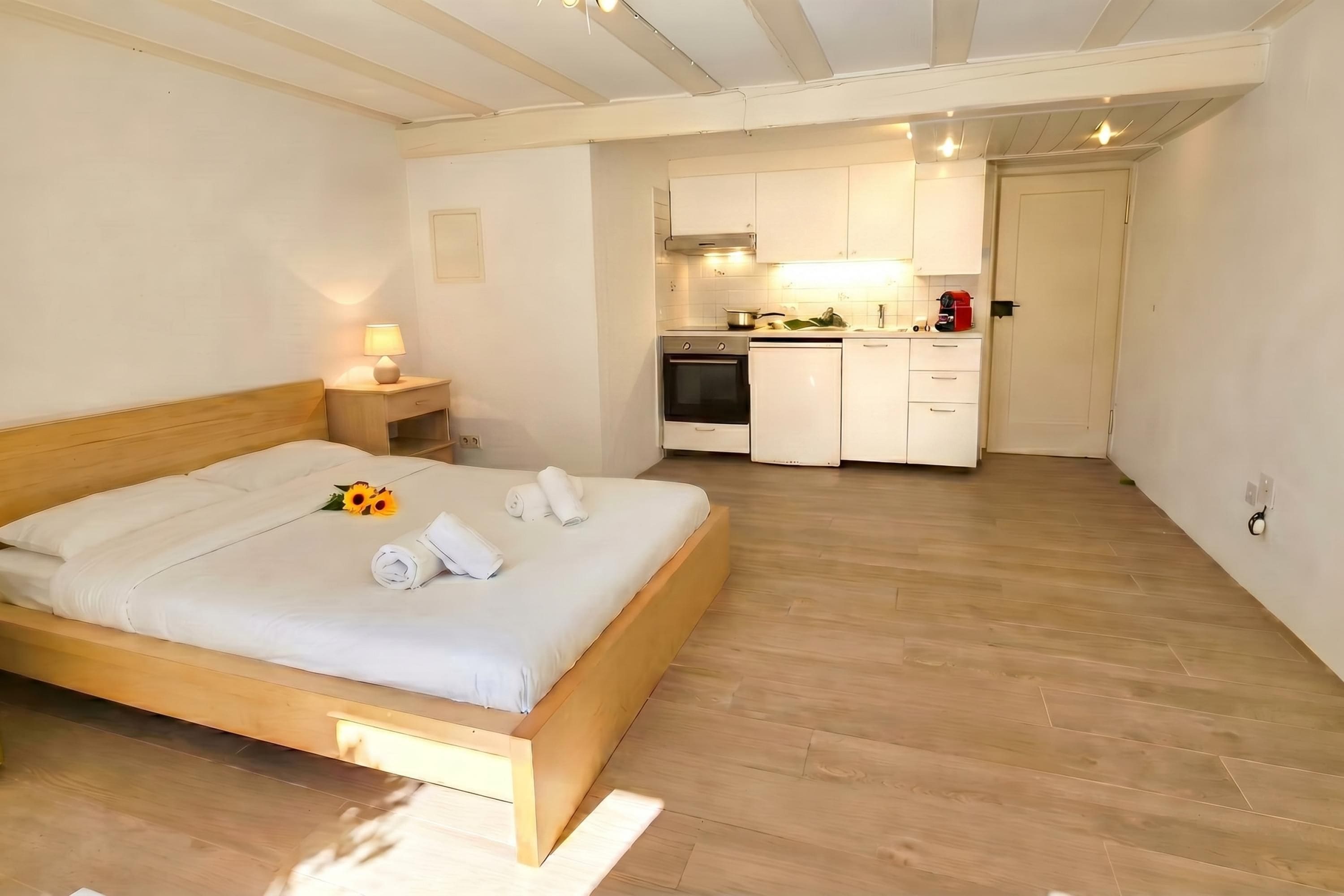 Ein Schlafzimmer mit einem hölzernen Bett, einem Nachttisch mit Lampe und einer Küchenzeile im Hintergrund.
