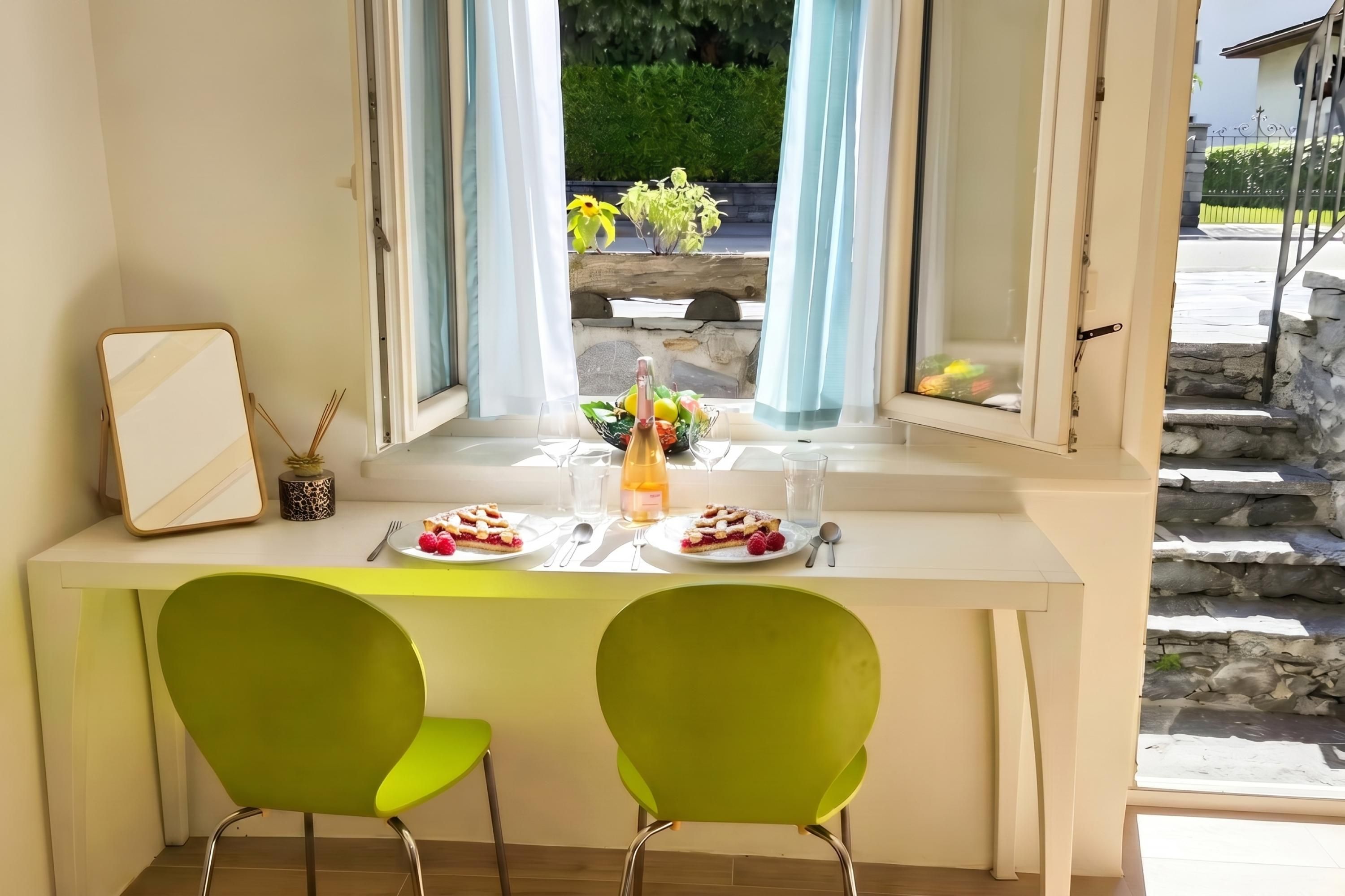 A dining area with a table set for two, a window with curtains, and a view of greenery outside.