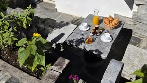 Una mesa de piedra con mantel blanco, una cesta de cruasanes, una jarra de jugo de naranja y una taza con platillo.