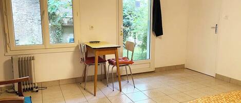 A room with a bed, a table, two chairs, a radiator, and a window with a view of greenery.