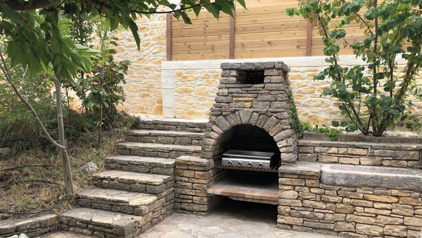 Cocina de piedra al aire libre con una parrilla, escalones que la conducen y una zona de estar con cojines.