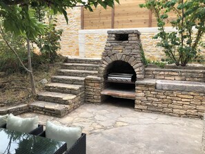 A stone-built outdoor kitchen with a grill, steps leading up to it, and a seating area with cushions. - Charming house that sleeps 10—luxury—with a pool and air conditioning (Barjols)