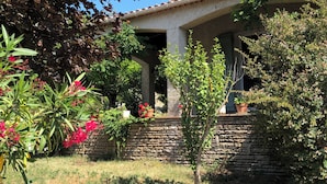 Un edificio de dos pisos con fachada de piedra, un porche cubierto y un jardín con diversas plantas y flores.