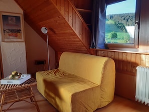 Een kamer met een gele bank, een rieten tafeltje met appels, een lamp en een raam met uitzicht op een bos.