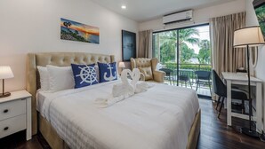 A hotel room with a bed, a desk, a chair, a nightstand, and a view of the outdoors. - Beachside 1BR•Walk to Rawai•Cozy-title rawai C201 (ตำบล ราไวย์)
