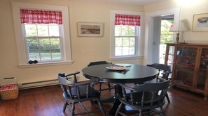 Un comedor con una mesa redonda y cuatro sillas, dos ventanas con cortinas rojas y un aparador con puertas de vidrio.