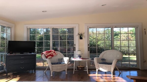 A room with a television, two wicker chairs, a small table, and large windows.