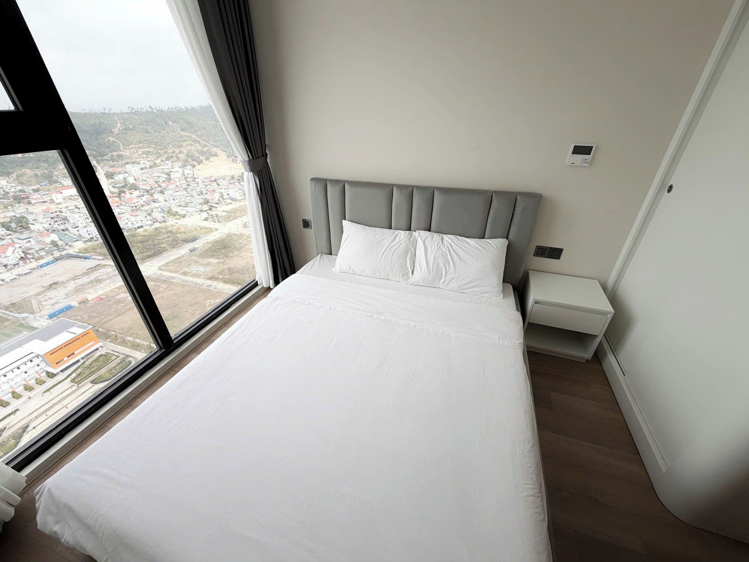 A hotel room with a large bed, a nightstand, a wall-mounted thermostat, and a view of a cityscape through a window.