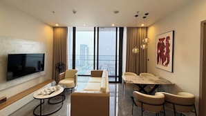 A modern living room with a flat-screen TV, a sofa, a dining table with chairs, and a large window with a city view. - Apartment in Icon 40 Ha Long   (Bai Chay)