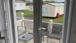 Una habitación con una puerta corrediza de vidrio que da a una terraza exterior con mesa y sillas, vista hacia una casa móvil y un área con césped.