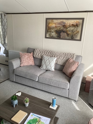 A grey sofa with patterned cushions, a framed abstract painting, a radiator, and a small table with a notebook, a map, and a can. - Lodge Sleeps 4, 2 Beds 2 Bathrooms (Morpeth)