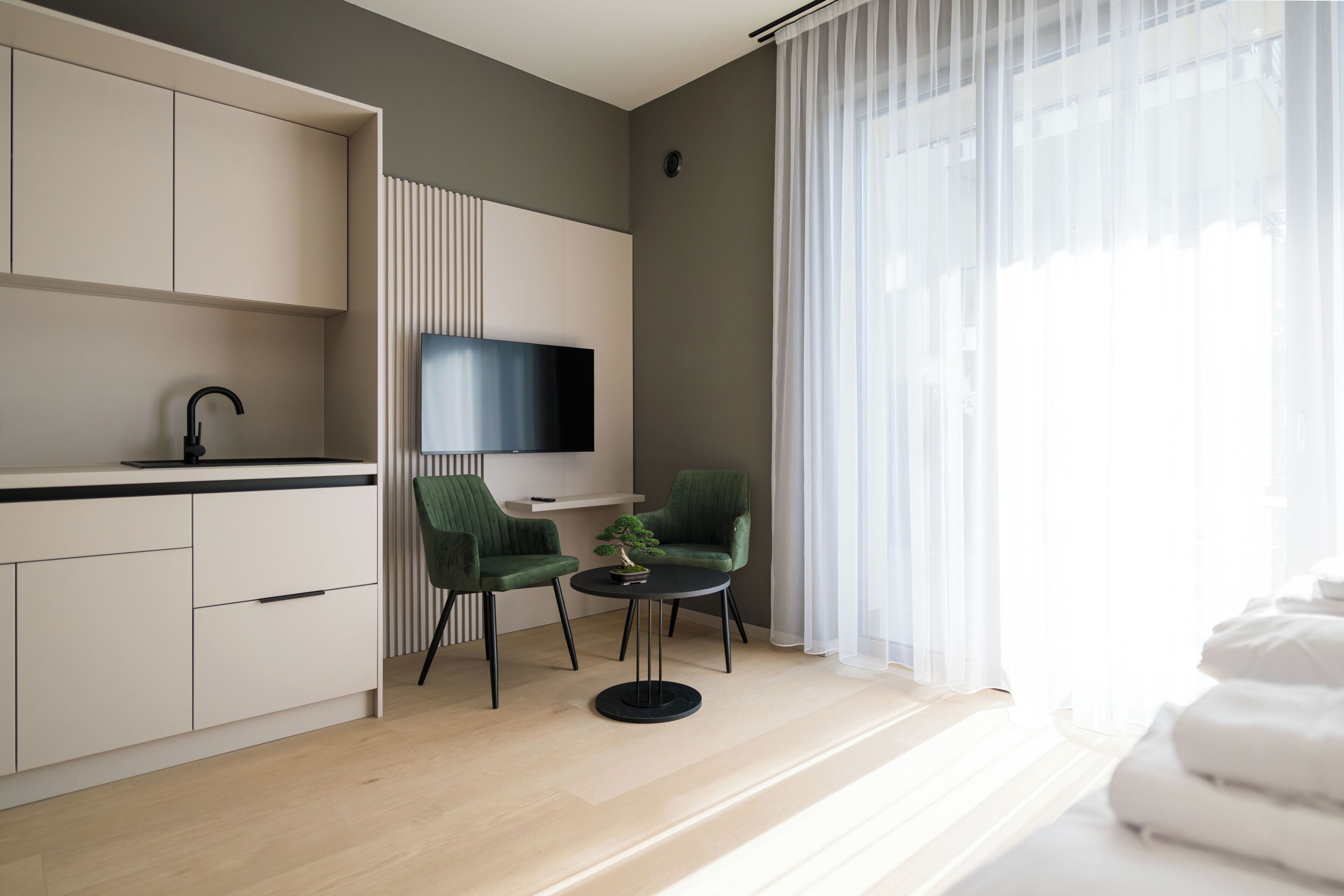 A modern hotel room with a kitchen area, a flat-screen TV, two green chairs, a small round table, and a bed with white linens.