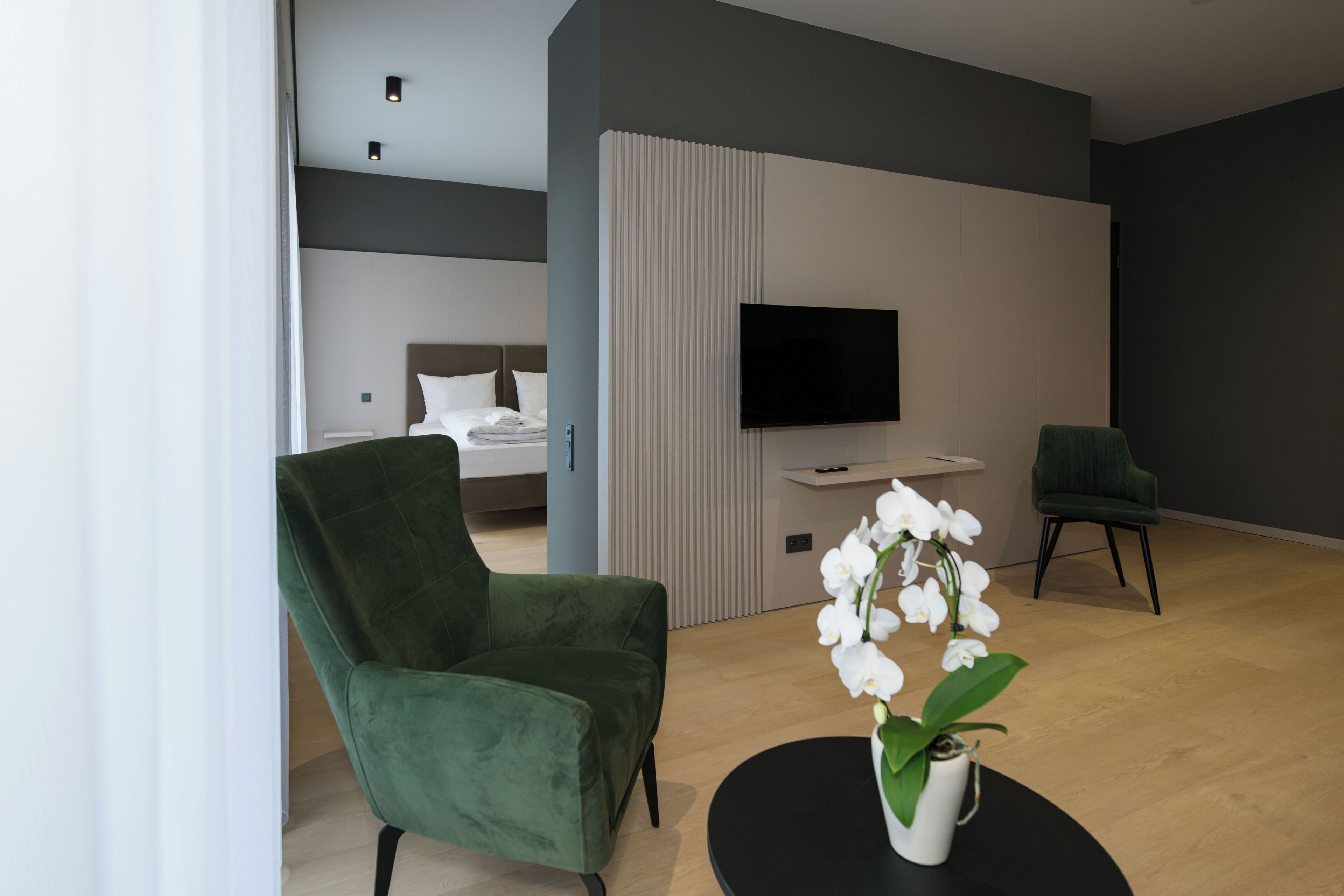 Moderni hotellihuone, jossa on vihreä nojatuoli, pyöreä musta pöytä, jonka päällä on valkoinen orkidea, seinälle asennettu litteänäyttöinen televisio ja valkoisilla lakanoiden päällä oleva sänky.