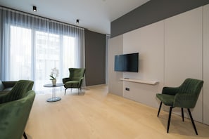 Ein modernes Wohnzimmer mit einem Wand-Fernseher, einem runden Tisch mit Topfpflanze und zwei grünen Sesseln.