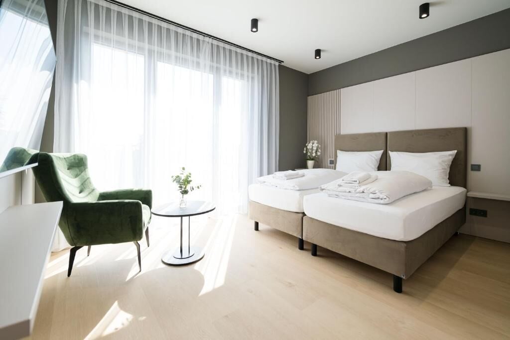 Une chambre d’hôtel moderne avec un lit, un fauteuil vert, une petite table avec une plante et une vue sur l’extérieur à travers des rideaux transparents.