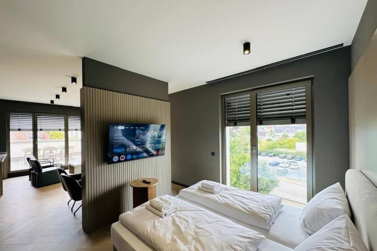A modern hotel room with a bed, a TV, a window with blinds, and a view of the outside.