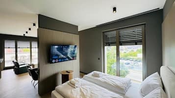A modern hotel room with a bed, a TV, a window with blinds, and a view of the outside.