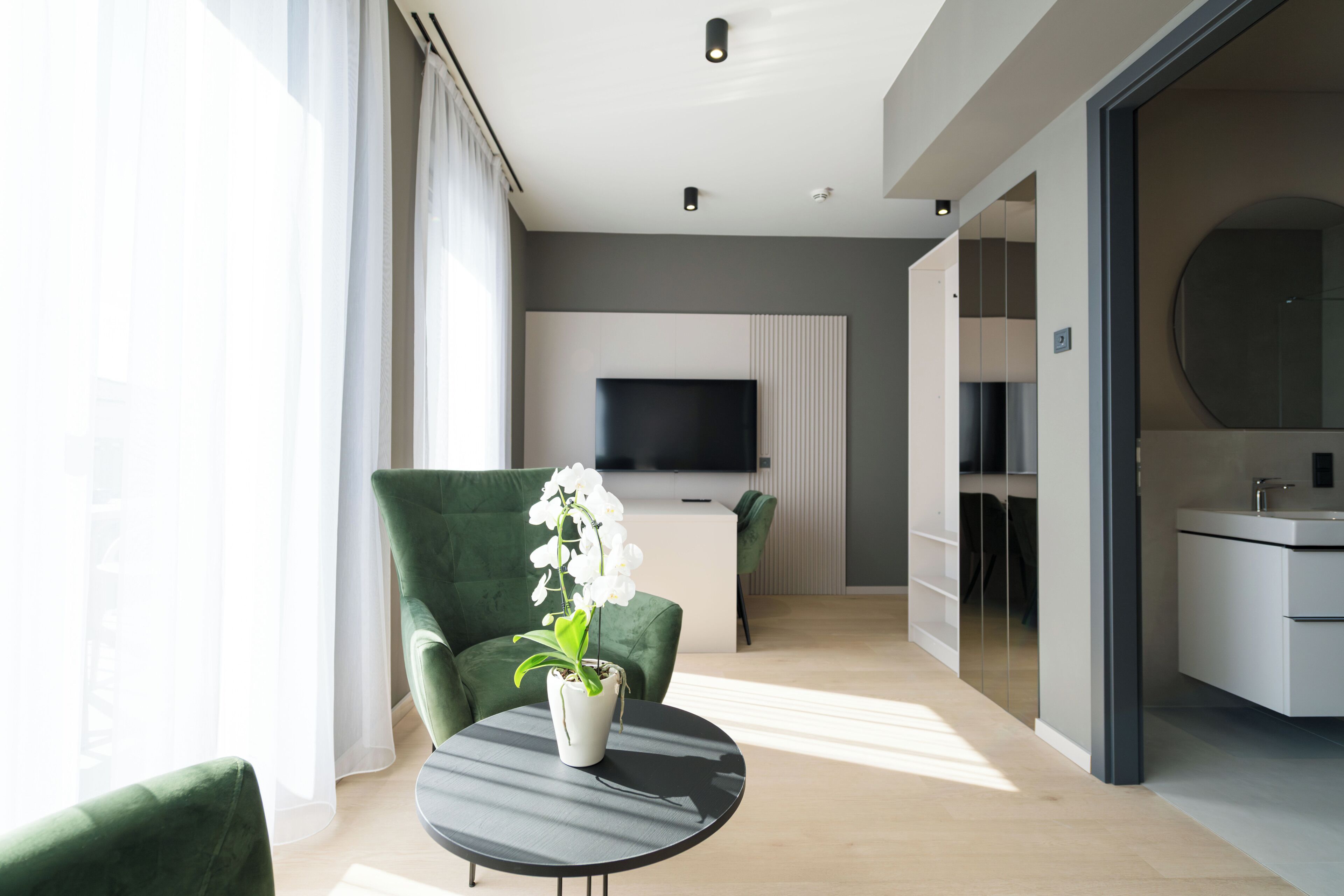 Une chambre d’hôtel moderne avec un fauteuil vert, une table ronde ornée d’une orchidée en pot, un téléviseur à écran plat et une salle de bain au design minimaliste.