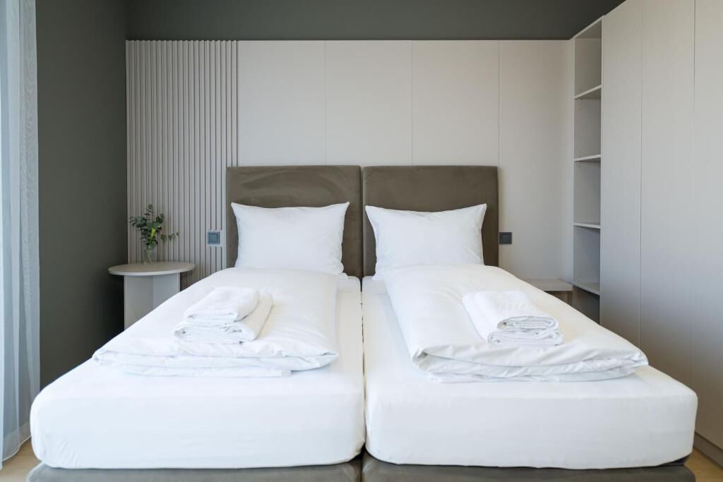 A double bed with white linens and towels, a bedside table with a vase, and a vertical panel on the wall.