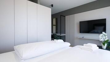 A modern hotel room with a bed, a flat-screen TV mounted on the wall, a small desk, and a potted orchid.