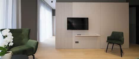 A modern living room with a flat-screen TV mounted on a wall, two green armchairs, a small side table with a vase of white flowers, and a light-colored wooden floor.