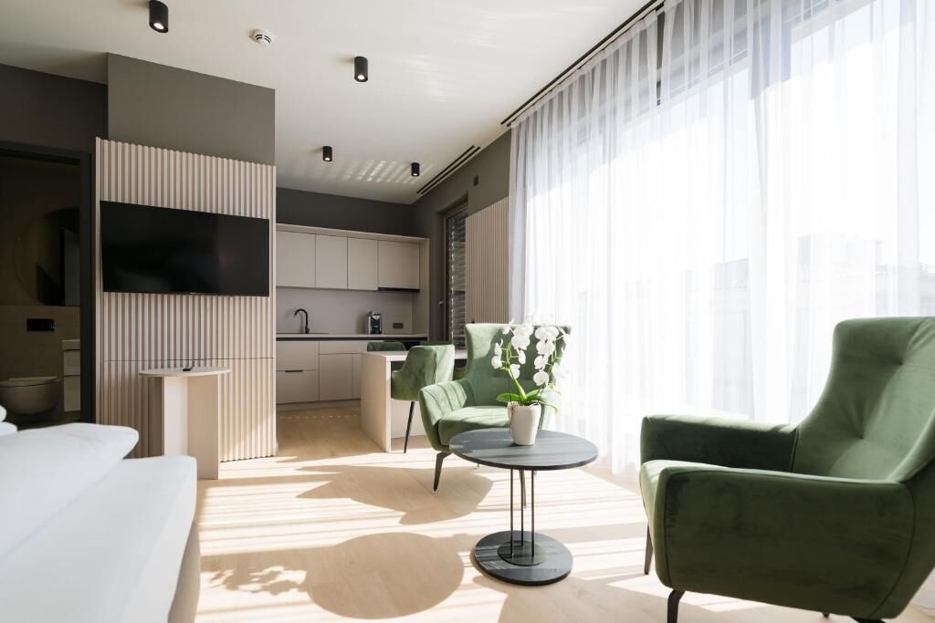 A modern hotel room with a sofa, a green armchair, a small round table with a potted orchid, a built-in TV, and a kitchen area visible through an open door.