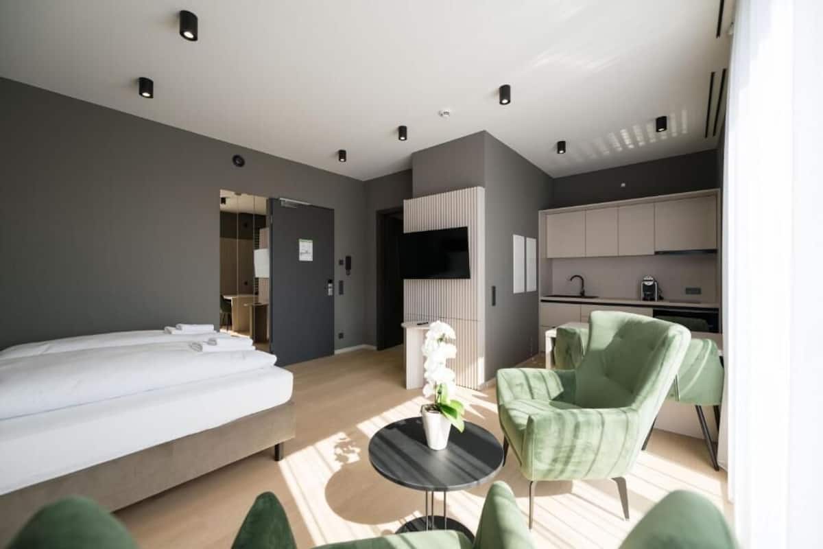 A modern hotel room with a bed, a small round table with a plant, green armchairs, a kitchenette, and a bathroom visible through an open door.