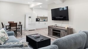 A modern living room with a flat-screen TV, a sofa, a coffee table, and a kitchen area in the background. - Dilalla Residence -The Hidden Vine Retreat (Niagara Falls)