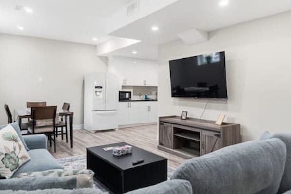 A modern living room with a flat-screen TV, a sofa, a coffee table, and a kitchen area in the background.