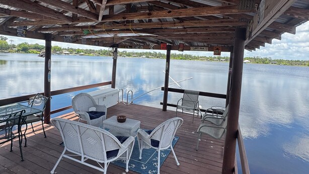 A covered outdoor deck with a table and chairs, overlooking a calm body of water. - Cozy 2-bedroom cottage in charming Lake Placid with AC,and breathtaking sunsets (Lake Placid)