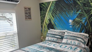 A bedroom with a bed featuring a tropical-themed comforter, a wall-mounted air conditioner, and a window with blinds. - Cozy 2-bedroom cottage in charming Lake Placid with AC,and breathtaking sunsets (Lake Placid)