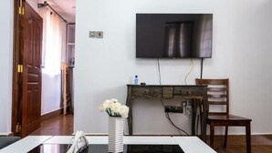 Una habitación con una televisión de pantalla plana montada en la pared, un escritorio de madera con una silla, un jarrón con flores blancas sobre una mesa y una puerta que da a otra estancia.