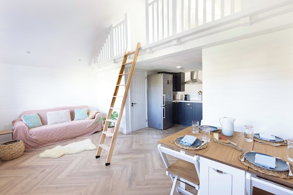 Un espace de vie de style « loft » comprenant une table à manger en bois pour quatre personnes, une cuisine équipée d’appareils modernes, un lit en mezzanine et une échelle.