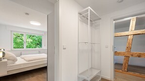 Una habitación con un estante blanco, una cama con cabecera blanca y una ventana que da a follaje verde.