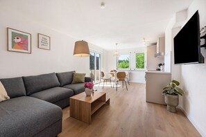Ein modernes Wohnzimmer mit einem grauen Sofa, einem Holztisch und einem Blumenstrauß. Es gibt einen Essbereich mit Tisch und Stühlen sowie eine Küche mit an der Wand montiertem Fernseher.