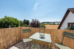 Ein Außenbereich mit Tisch und Stühlen, einem Bambuszaun und Blick auf ein Feld und Hügel im Hintergrund.
