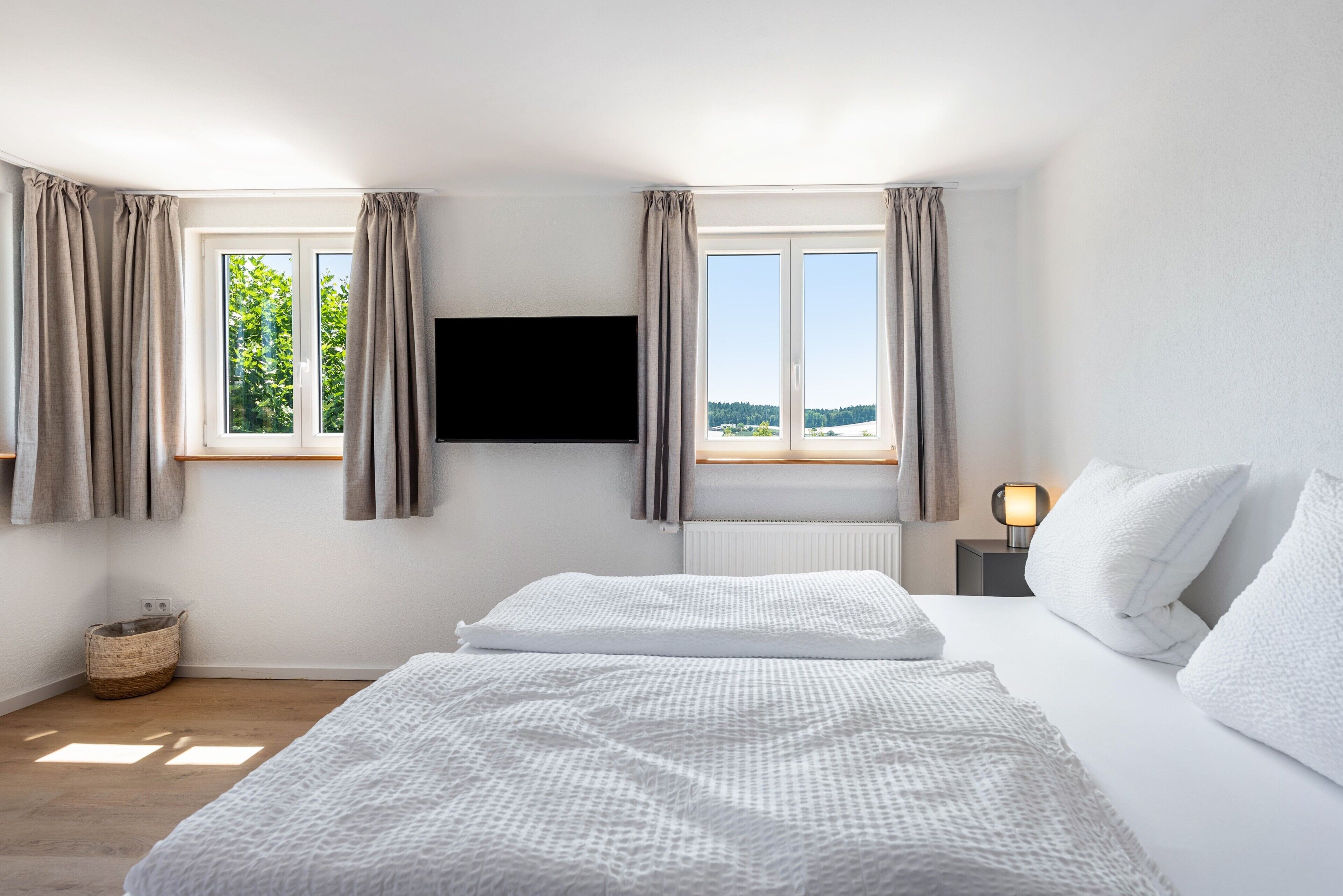 Een slaapkamer met een bed, een aan de muur bevestigde televisie, een raam met gordijnen, een nachtkastje met een lamp en uitzicht op groen buiten.
