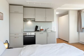 A compact kitchen with a stove, oven, and sink, a bed with a white blanket, and a window with curtains. - Apartment Mainau with Private Kitchen (Salem)