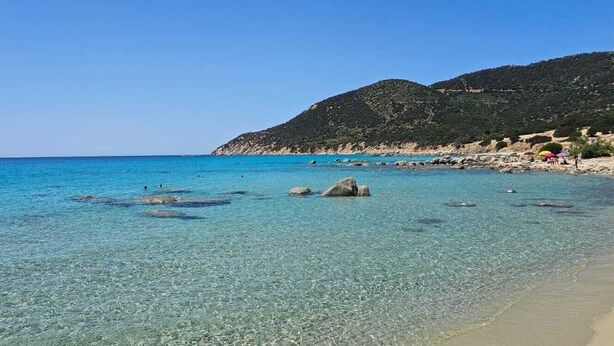 Una playa con agua cristalina, rocas y un paisaje montañoso al fondo.