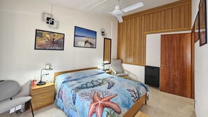 Un dormitorio con cama, escritorio, silla, armario, ventilador de techo y dos cuadros en la pared.