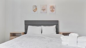 Una cama bien hecha con ropa blanca, un cabecero gris y una mesita de noche con una toalla blanca doblada.