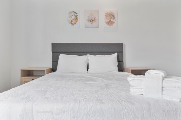 Una cama bien hecha con ropa blanca, un cabecero gris y una mesita de noche con una toalla blanca doblada.