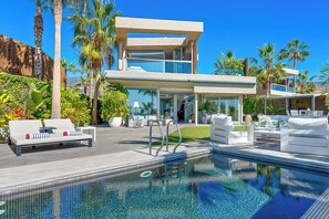 Une maison moderne à deux étages, dotée d’une piscine, de mobilier d’extérieur et d’une végétation luxuriante.