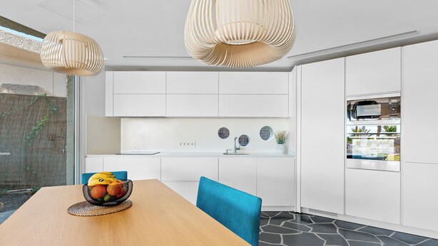Une cuisine moderne avec des meubles blancs, une table à manger sur laquelle se trouve un panier de fruits, et une chaise bleue.