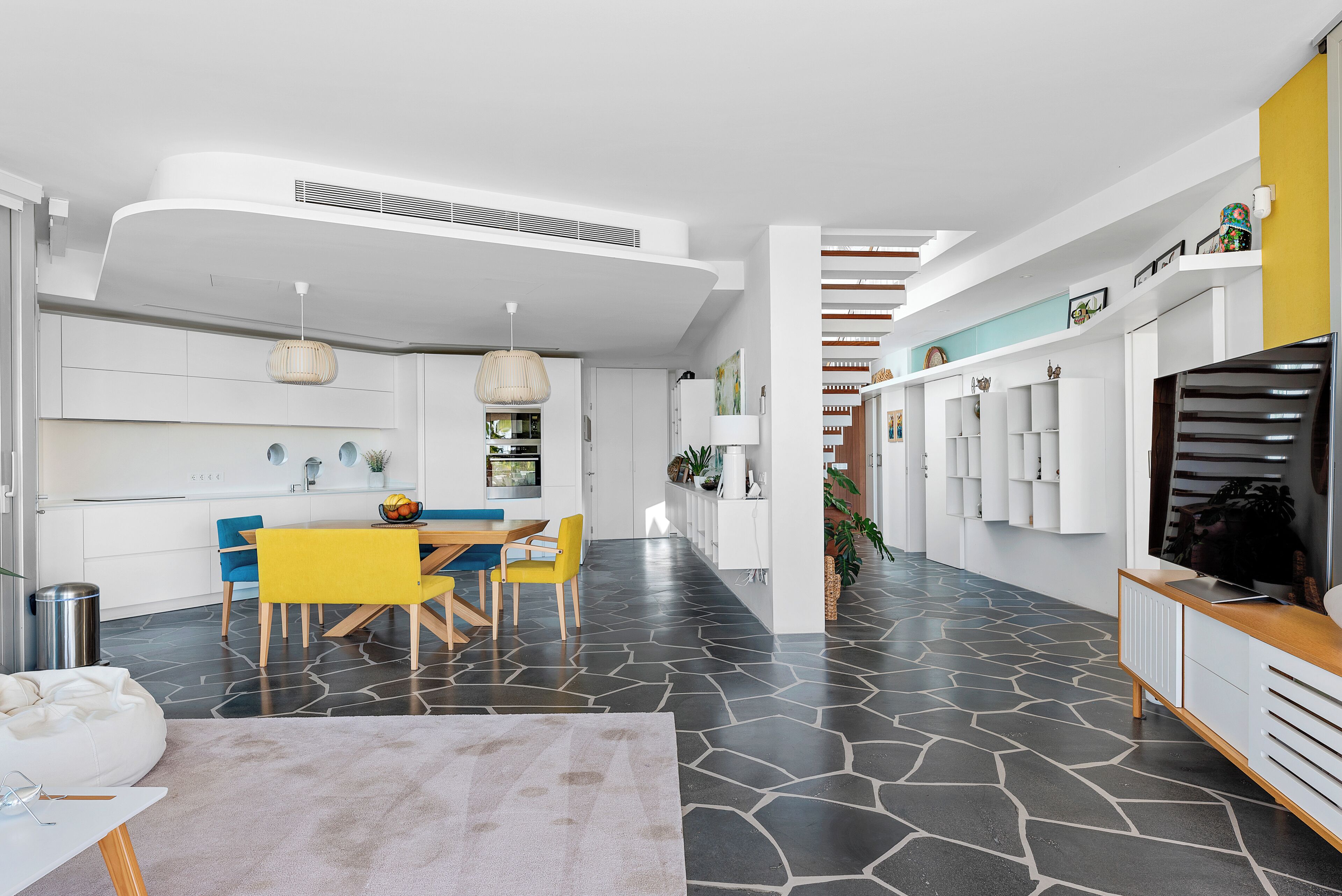 Une cuisine moderne avec des meubles blancs, un coin repas avec des chaises jaunes et un escalier menant à une mezzanine.