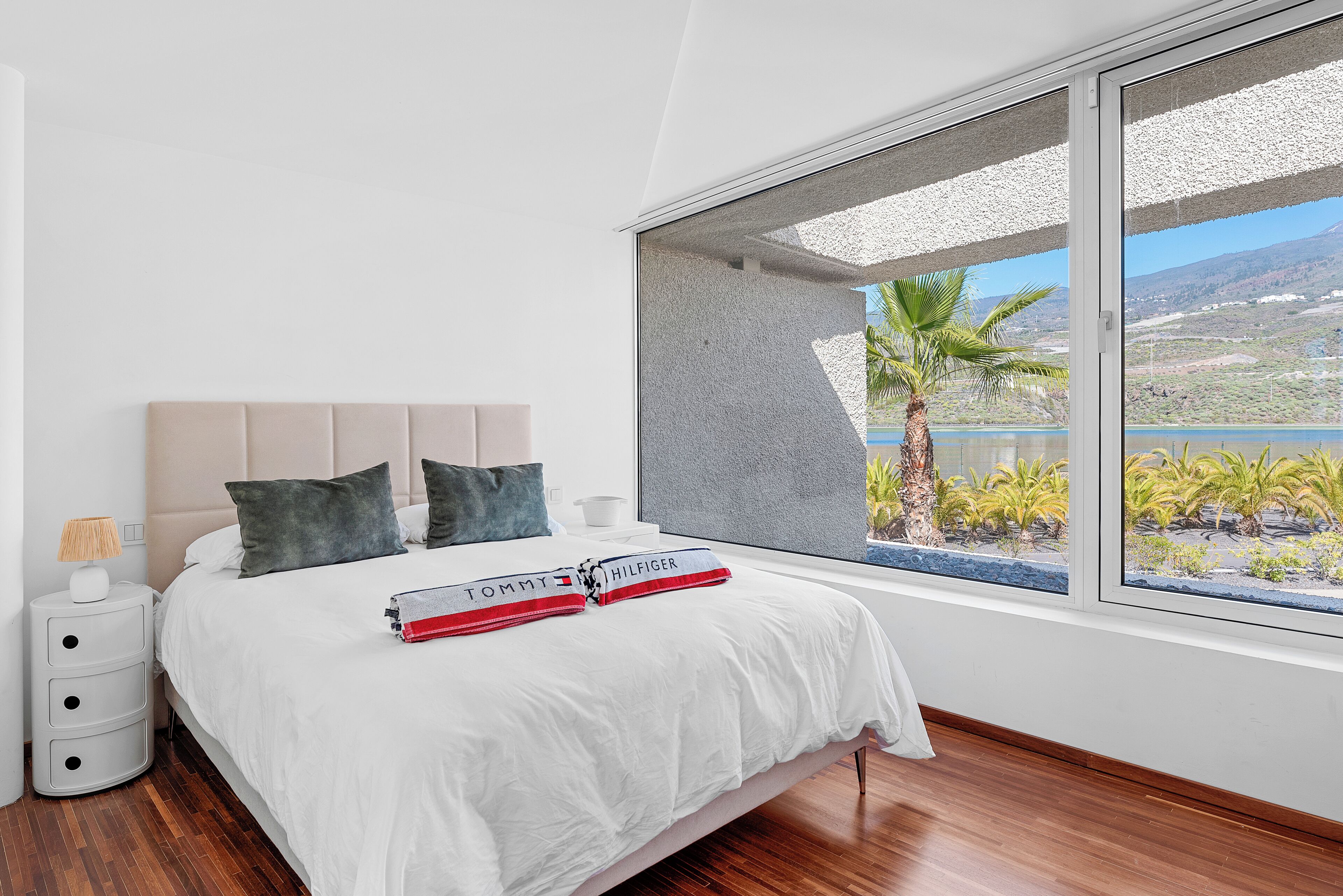 Een slaapkamer met een groot raam, een bed met witte beddengoed, een nachtkastje en een lamp.